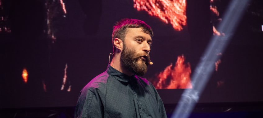 a bearded man talking to public with a microphone