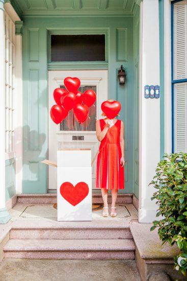 a girl covering her face with red heart shaped baloon standing next to the box pf baloons