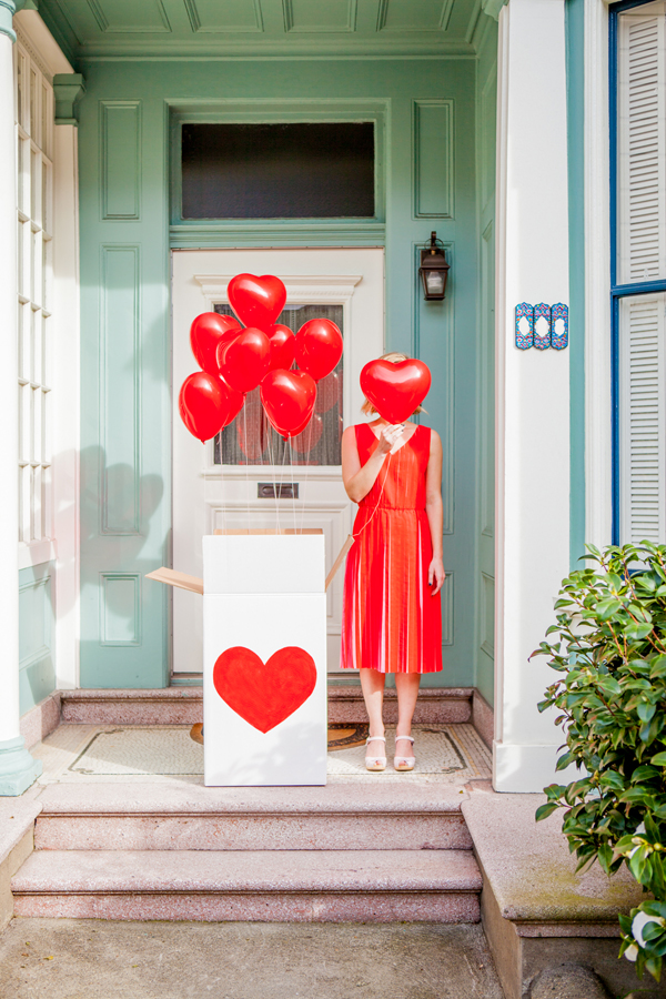 a girl covering her face with red heart-shaped baloon standing next to the box pf baloons