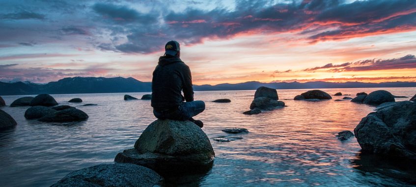 a guy sitting on the rock in the sea at sunset