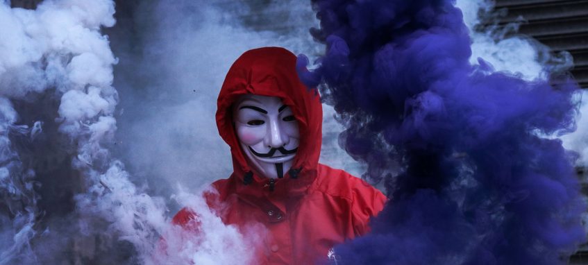 a guy wearing an anonymous mask stands in clouds of smoke