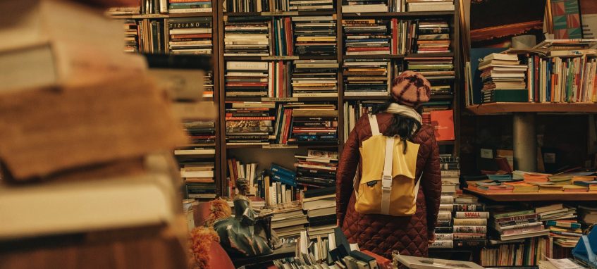 a student with a backpack standing in a library surronded by a pile of books