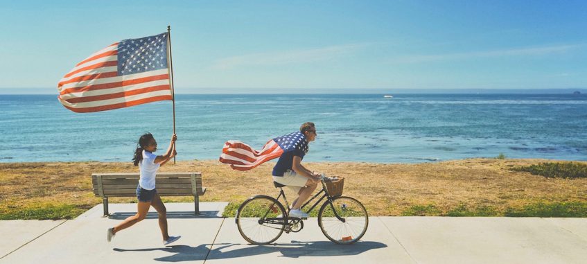 the girl with American flag is chasing the guy on bicycle wrapped with the flag of United States